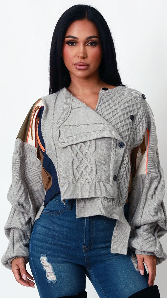 A woman modeling a light gray cable-knit cropped jacket with mixed-color panels in beige, brown, and navy, paired with blue jeans against a plain white studio background.