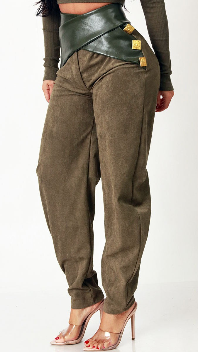 A model is shown from the waist down wearing high-waisted, baggy olive-brown pants with a wide dark green wrap-style belt and gold-tone hardware, paired with clear high-heeled sandals.