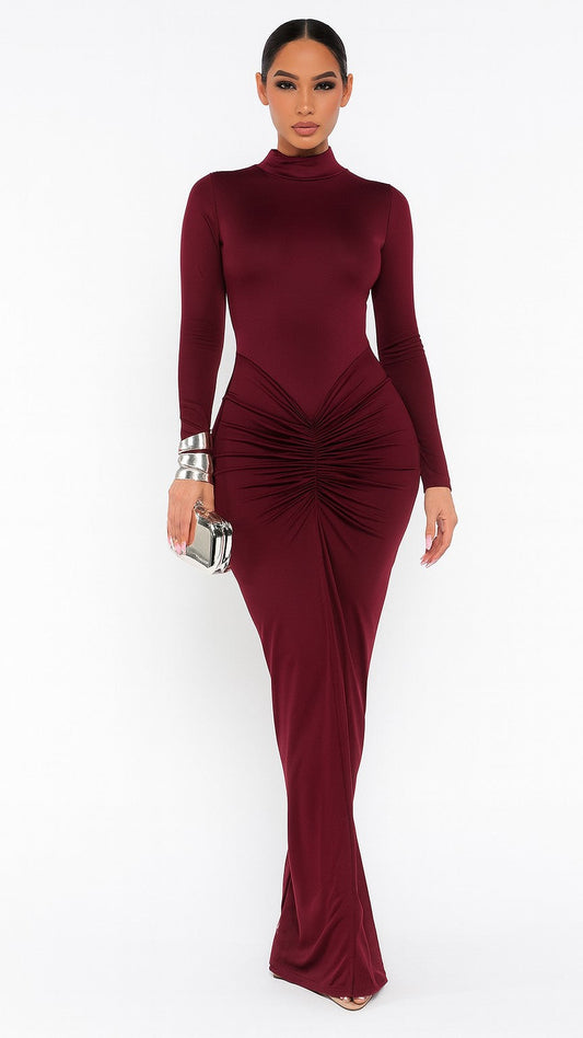 A woman modeling a long-sleeve, high-neck, bodycon maxi dress in deep burgundy with a ruched gathered detail at the waist, set against a plain white studio background.