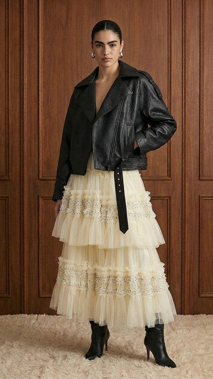 A model stands against a dark wood-paneled wall wearing a black leather moto jacket over a cream tulle tiered skirt with lace trim, paired with black ankle boots.