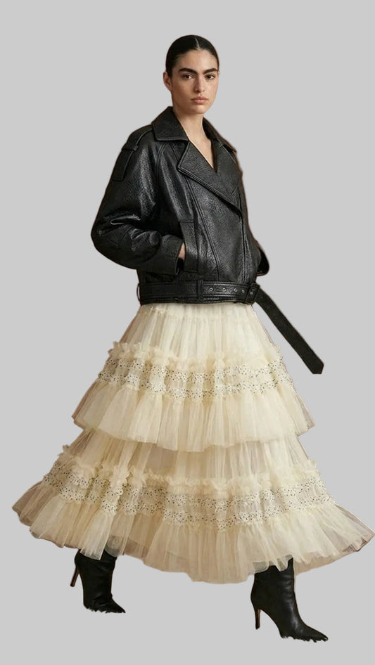 A model stands in front of a dark wood paneled wall wearing a black leather moto jacket with a black belt, paired with a voluminous cream tulle tiered skirt with lace trim, and black heeled ankle boots.