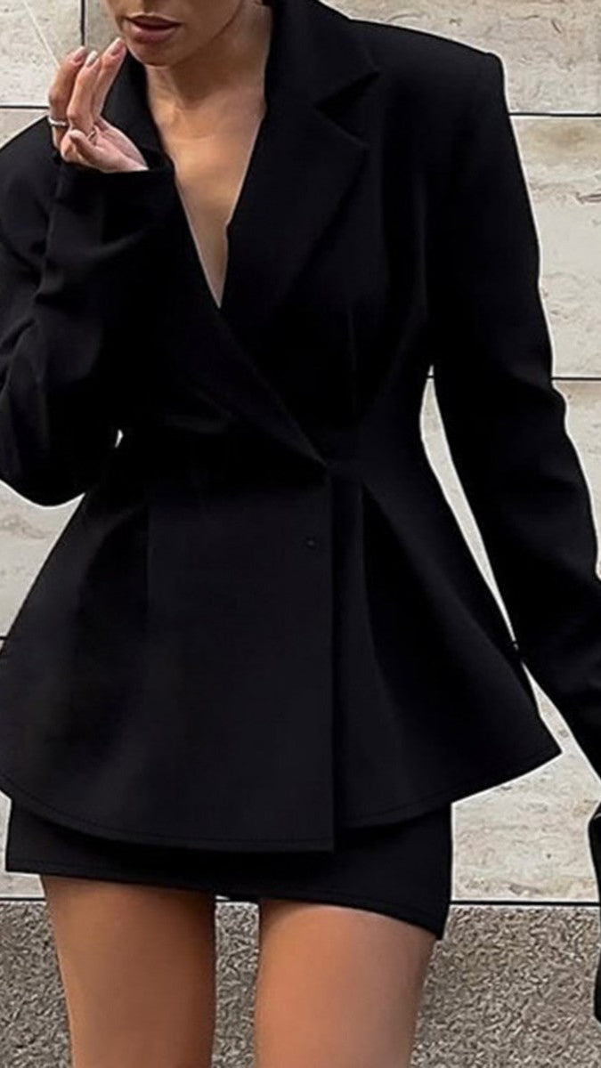 A woman wearing a black tailored blazer-style mini skirt with structured shoulders and a wrap-front silhouette, paired with a matching black mini skirt, standing against a light stone wall.