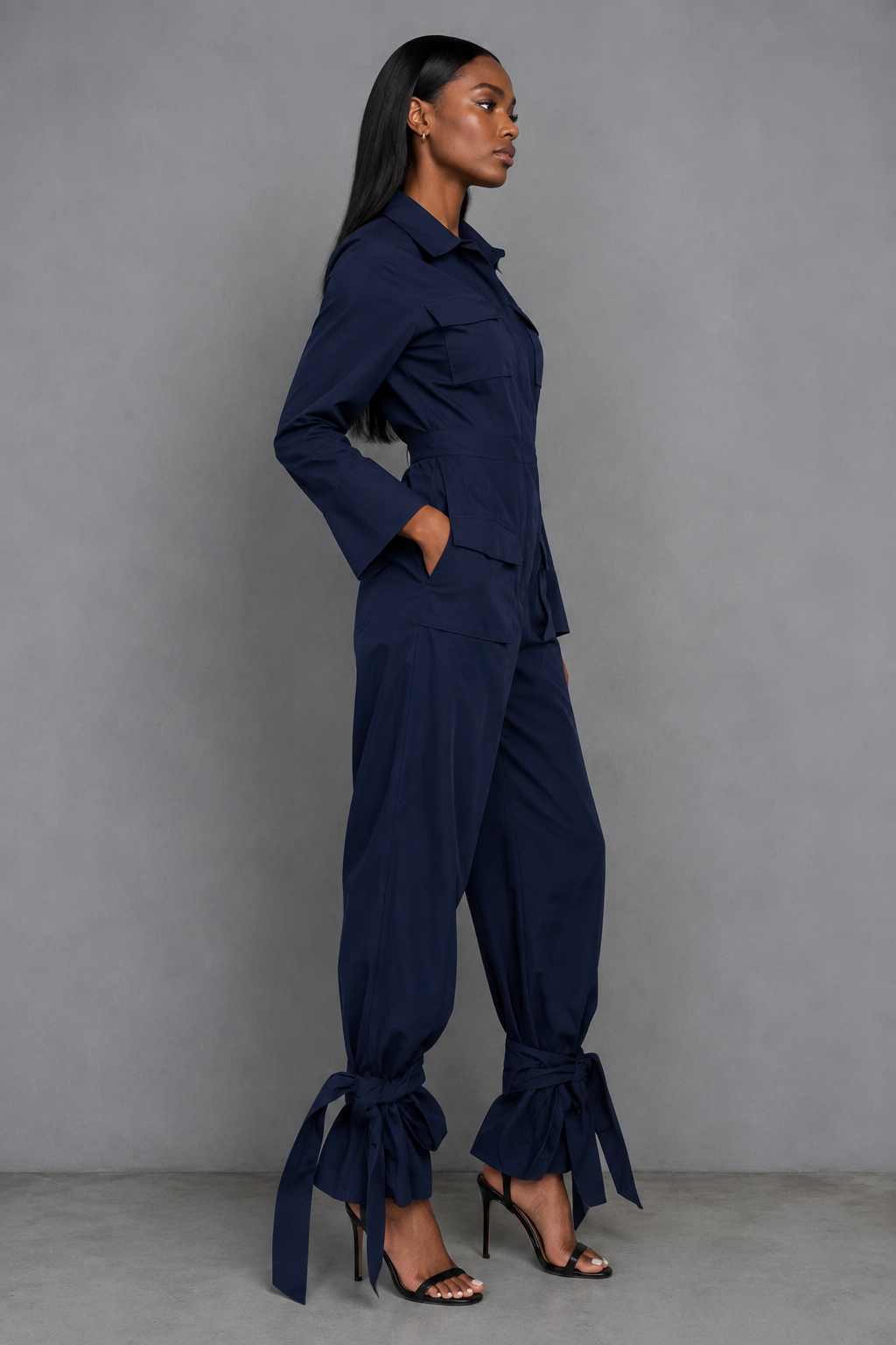 A woman modeling a navy blue long-sleeve jumpsuit with gathered balloon-style legs and tie details at the ankles, posed against a neutral gray studio backdrop.