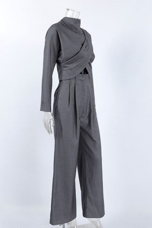 A dark gray long-sleeve jumpsuit displayed on a mannequin against a light gray studio background. The top features a high mock neck, dramatic diagonal wrap-style draping, and a cutout at the waist, paired with high-waisted, wide-leg trousers.