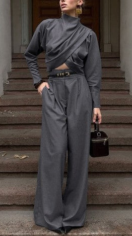 A model stands on stone stairs wearing a dark gray long-sleeve wrap-style crop top and matching wide-leg trousers, accessorized with a black belt, gold earrings, and a small black handbag.