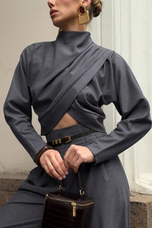 A model wearing a dark gray long-sleeve wrap-style top with a high mock neck and a matching gray skirt, holding a small dark brown handbag.