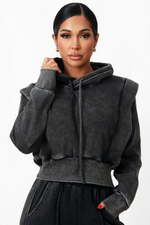 A woman modeling a dark gray cropped zip-up hoodie with wide shoulder panels and a drawstring hood, paired with black bottoms, against a plain white studio background.