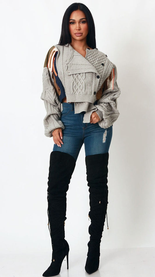 A woman modeling a light gray cropped knit jacket with brown and beige patterned shoulder panels, paired with blue jeans and black over-the-knee boots, against a plain white studio background.