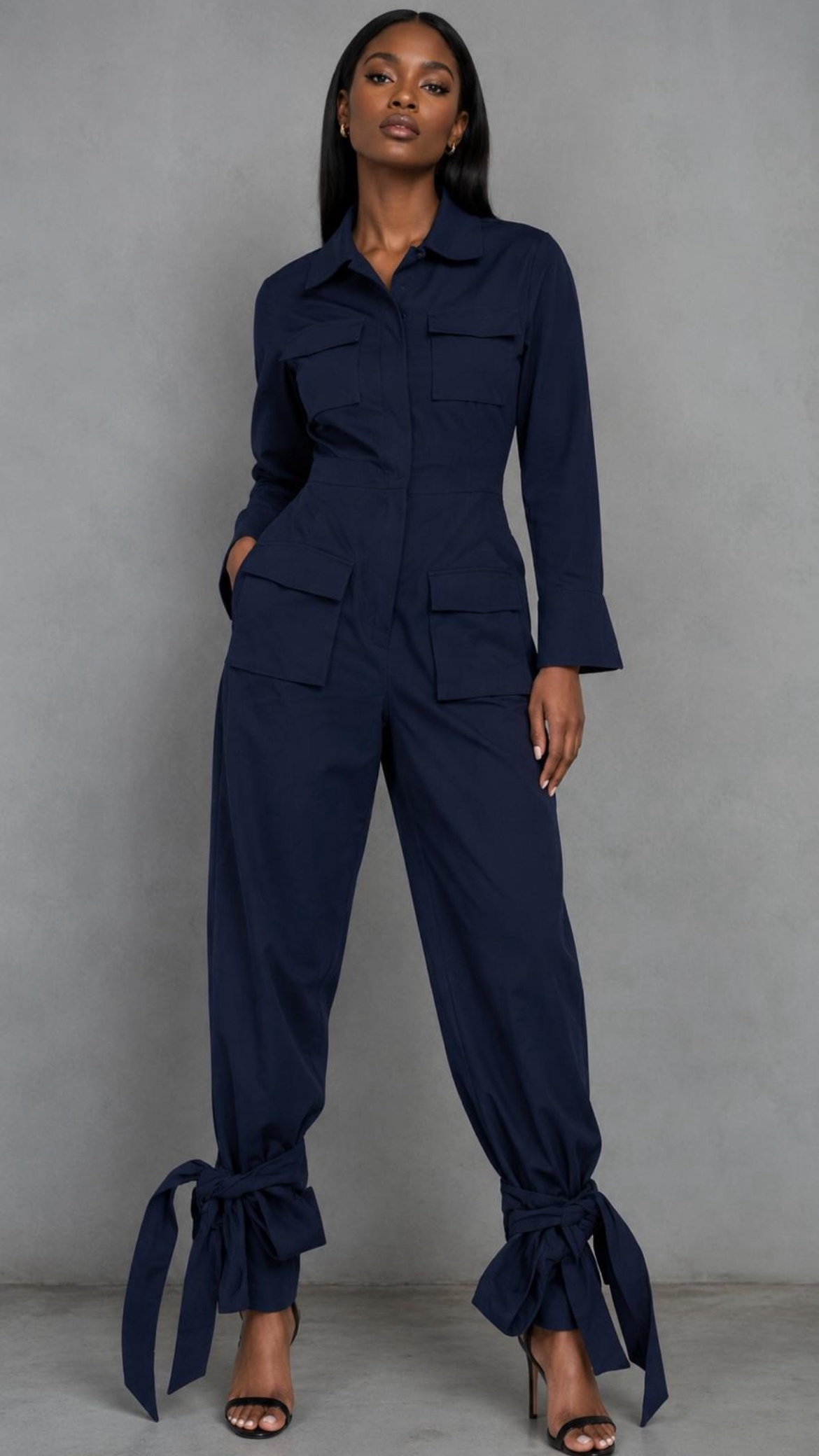 A woman modeling a navy blue long-sleeve cargo jumpsuit with front flap pockets and tie details at the ankles, standing against a plain gray studio background. Brand Materialguygal