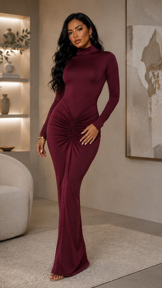A woman modeling a long-sleeve, high-neck, ruched floor-length dress in deep burgundy, standing in a modern, softly lit living room.