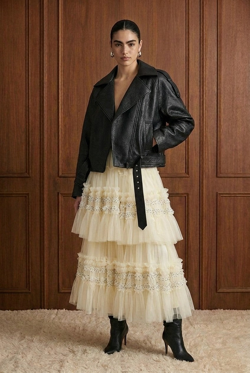 A model stands against a dark wood-paneled wall wearing a black leather moto jacket over a cream tulle tiered skirt with lace trim, paired with black ankle boots.