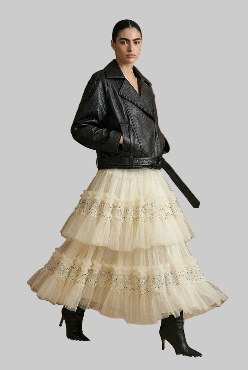 A model stands in front of a dark wood paneled wall wearing a black leather moto jacket with a black belt, paired with a voluminous cream tulle tiered skirt with lace trim, and black heeled ankle boots.