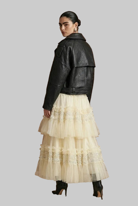 A model is shown from the back wearing a black leather jacket and a cream-colored tiered tulle skirt with lace details, standing in front of a dark wood paneled wall.