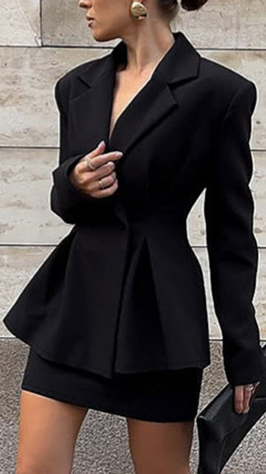A woman wearing a black tailored blazer with structured shoulders and a peplum waist, paired with a matching black mini skirt, standing against a light stone wall and holding a black clutch.