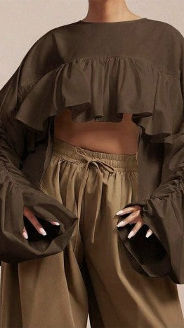 A model wearing an olive brown ruffled crop top with dramatic balloon sleeves and matching olive brown drawstring trousers, shown from the neck down against a neutral beige background.