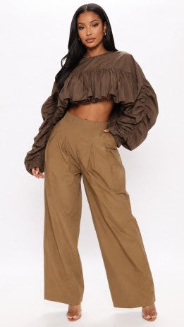 A model wearing a brown ruffled off-the-shoulder crop top and matching brown wide-leg trousers, standing against a plain white studio background.