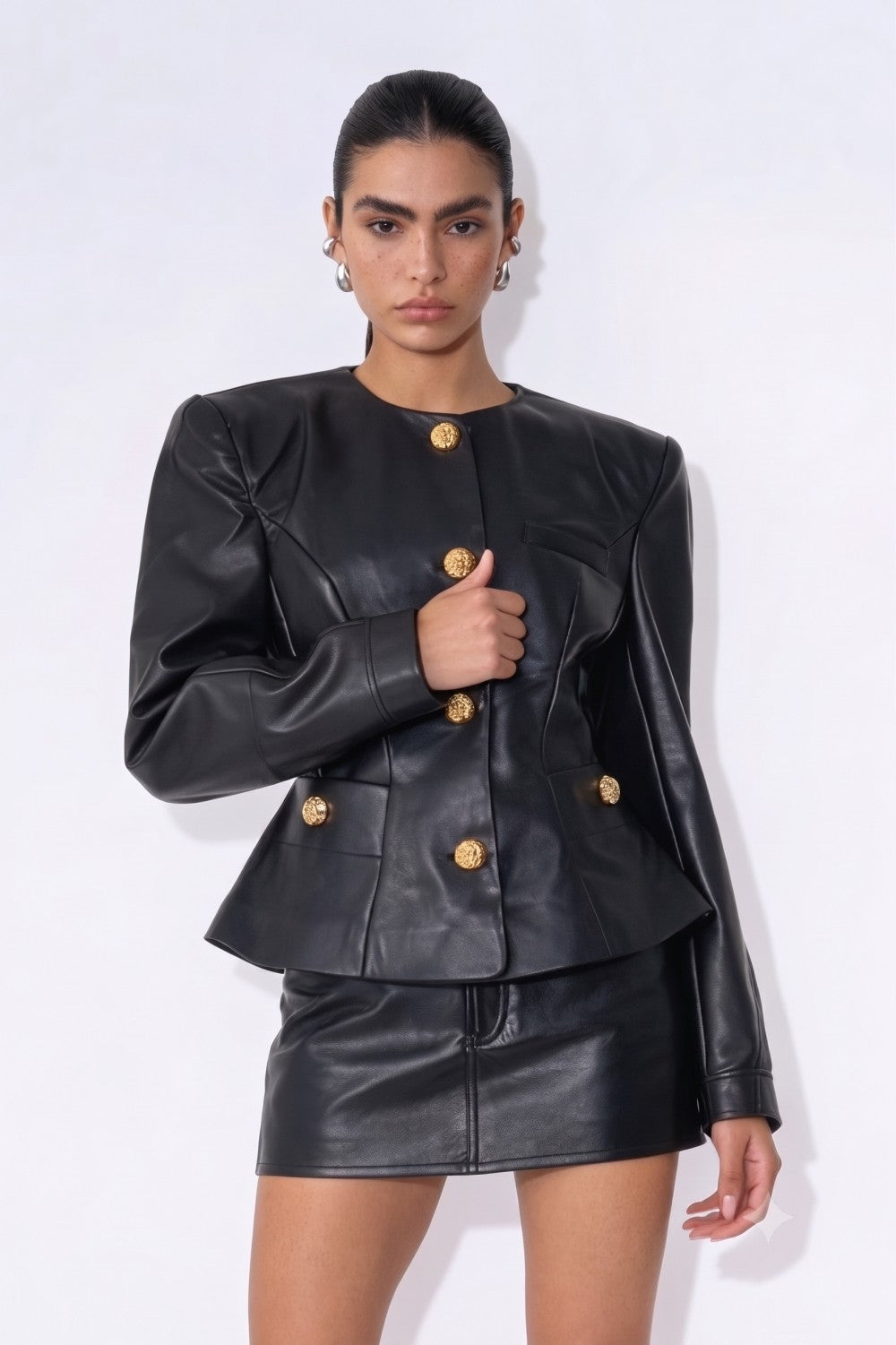 A model wearing a black faux leather peplum jacket and matching mini skirt set, featuring prominent gold-tone buttons, against a plain white studio background.